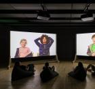 People watching large screens with projections of videos of children talking